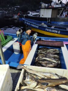 Fisher and his catch at night, with fishing boat in background, wearing orange overalls and a beanie. 