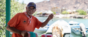 Photo of an older man in Mexico holding fishing line, wearing an orange shirt. You can see boats and water in the background, a bay, and hills behind him.