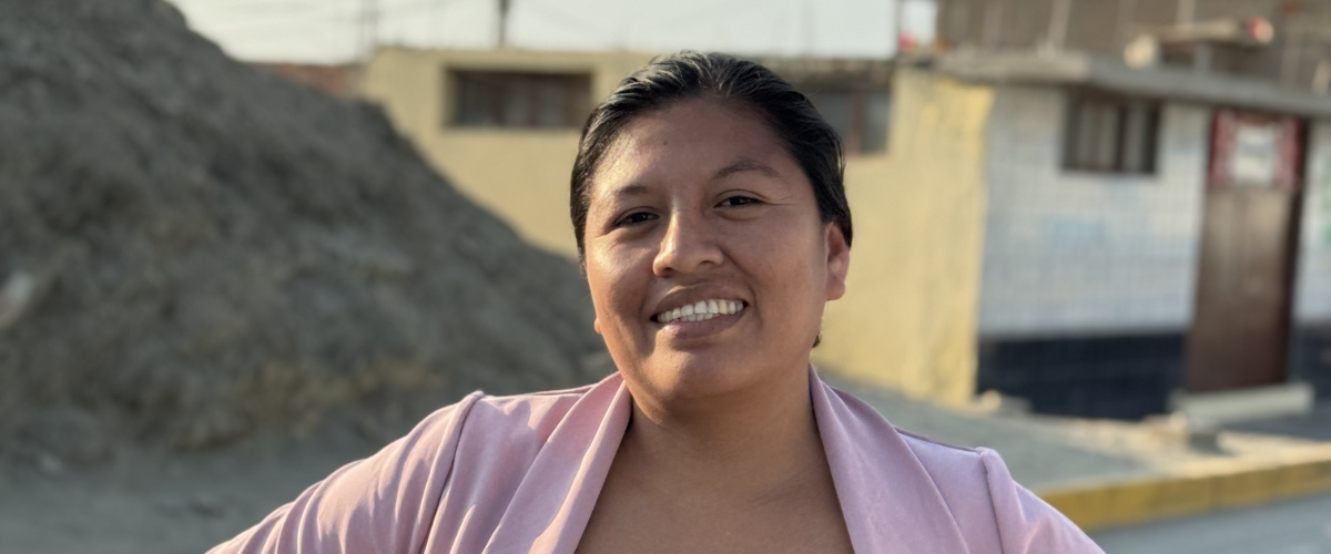 Photo of Gisella, woman standing on the street in her community in La Islilla Peru, with a house behind and mound of dirt.